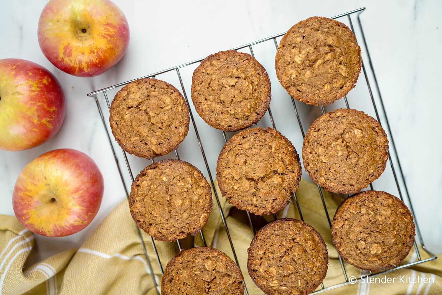 Applesauce Oatmeal Muffins Applesauce oatmeal muffins with oatmeal flecks on a baking rack with a yellow napkin and apples.