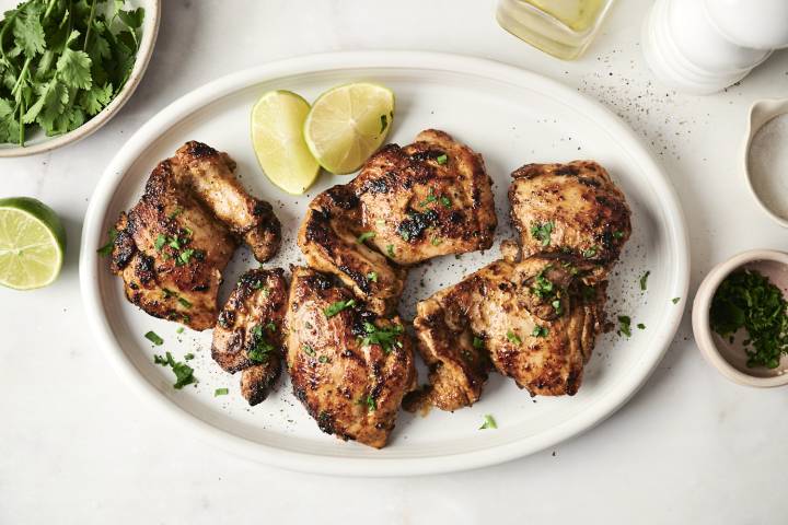 Juicy chipotle chicken thighs served on a white platter, garnished with fresh cilantro and lime wedges, surrounded by bowls of salt, herbs, and olive oil for a vibrant presentation.