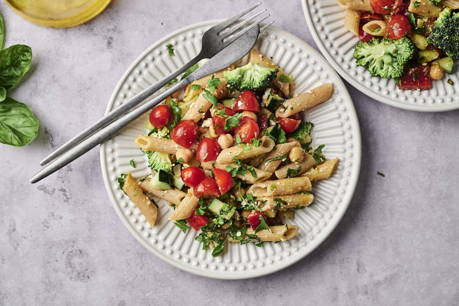 A vibrant plate of pasta mixed with cherry tomatoes, broccoli, and herbs, accompanied by a glass of olive oil and fresh basil leaves.