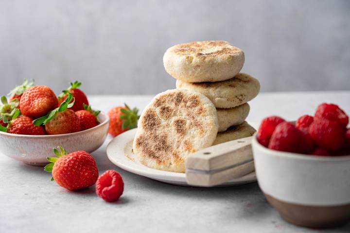 A plate with stacked English muffins and a cutting board on a gray surface. Nearby bowls hold fresh strawberries and raspberries, creating a fresh and inviting scene.