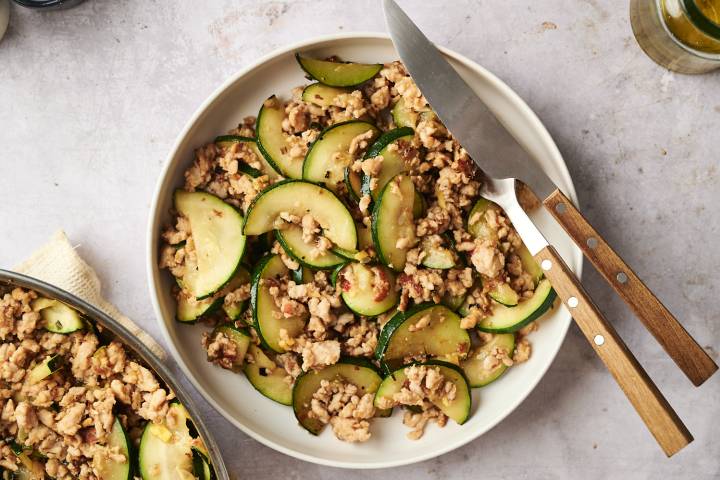 Bowl of ground turkey zucchini stir fry with tender zucchini slices and browned turkey, served with a knife and fork on the side.