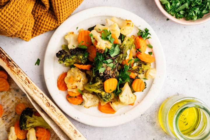 How to Roast Frozen Vegetables A white plate with roasted broccoli, cauliflower, and sliced carrots topped with fresh parsley. Surrounded by a yellow cloth, a dish of herbs, and olive oil.