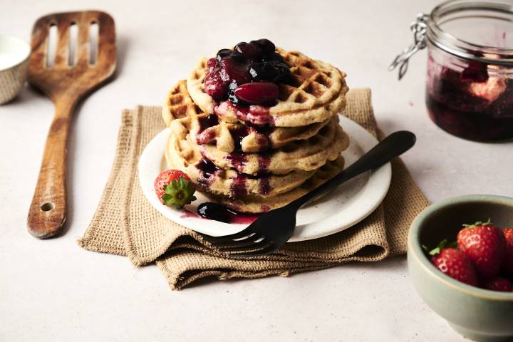 Low Carb Waffles Stack of golden low carb waffles topped with warm berry compote and syrup, served on a small plate with a black fork, fresh strawberries on the side, and a rustic jar of fruit sauce in the background.