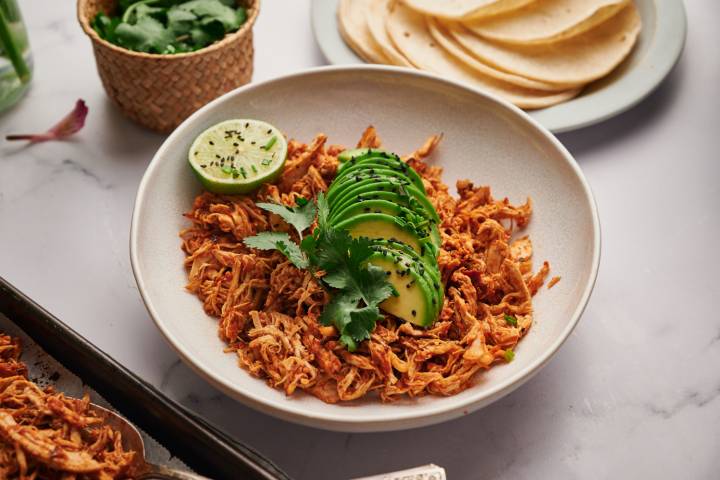 Easy Mexican Shredded Chicken (3 Ways!) Mexican shredded chicken with tomatoes, spices, and chipotle peppers in a bowl with avocado, lime, and corn tortillas.