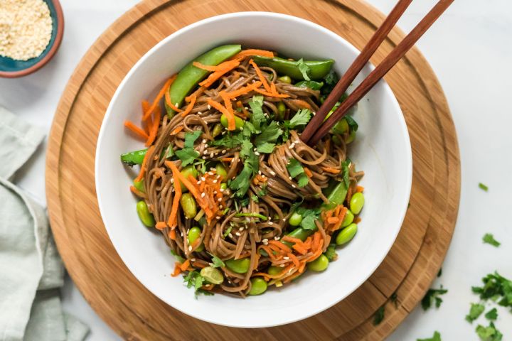 Asian Noodle Bowls Asian noodle bowls with soba noodles, sesame dressing, carrots, edamame, and snap peas in a white bowl with chopsticks.