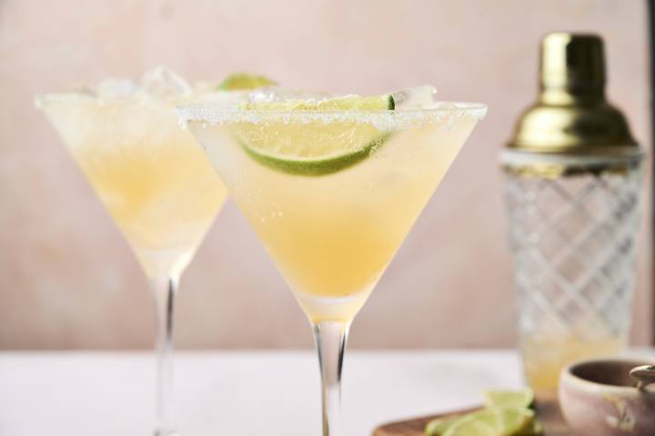 Skinny Margaritas Two chilled cocktails with lime slices sit beside a cocktail shaker, set against a soft pink background.