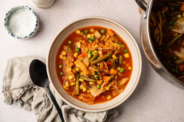 The Best Vegetable Soup Bowl of vibrant vegetable soup with corn, beans, carrots, and cabbage in tomato broth. Nearby, a black spoon on a beige napkin, with a salt container.