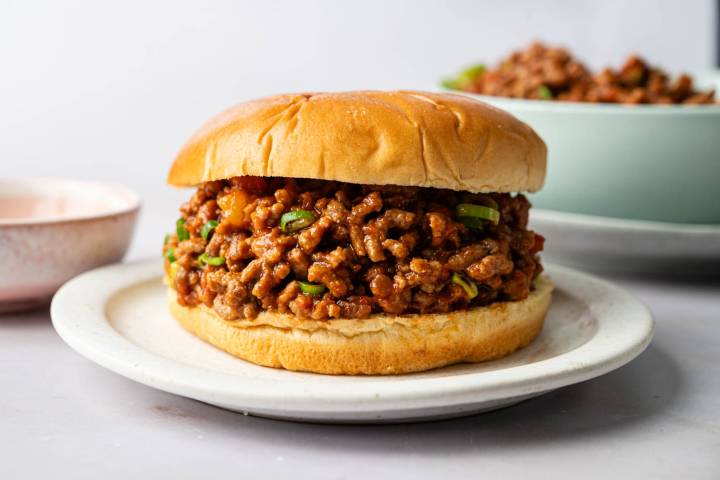 A juicy sloppy joe sandwich is centered on a white plate. The bun is golden brown, holding a savory, saucy ground meat filling. 