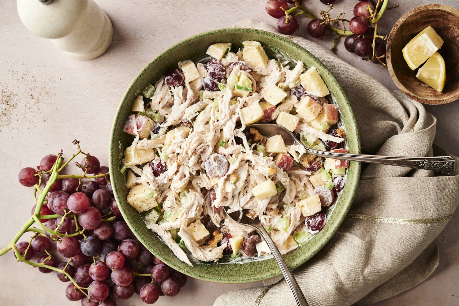 Overhead view of Waldorf chicken salad in a green bowl, made with shredded chicken, red grapes, celery, apples, and walnuts, served with two spoons.