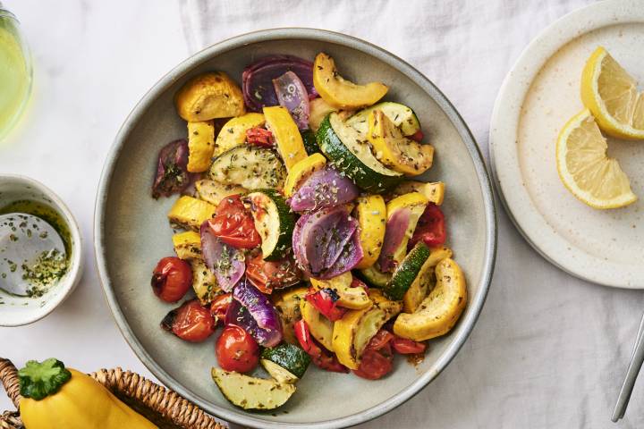 A bowl of colorful roasted vegetables, including zucchini, yellow squash, and cherry tomatoes, garnished with herbs. Lemon slices nearby.